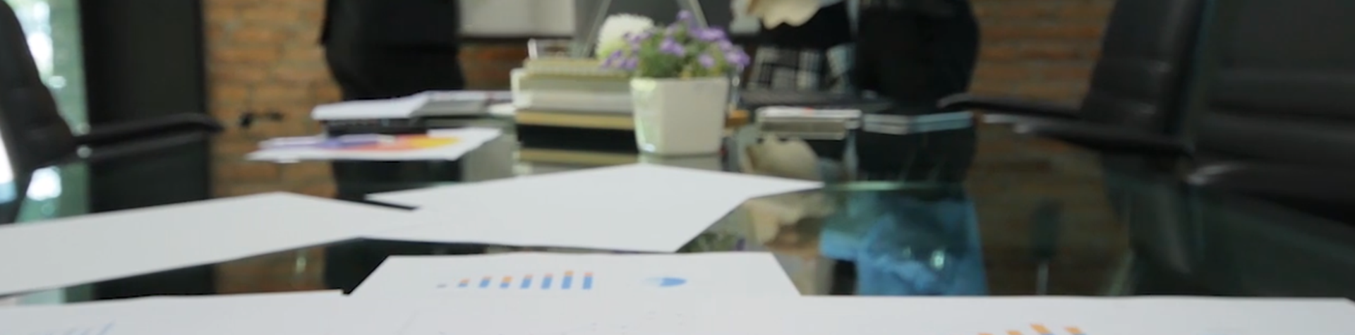 A boardroom of people have a discussion in front of a whiteboard, print outs of graphs on the table in front of them