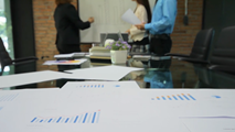 A boardroom of people have a discussion in front of a whiteboard, print outs of graphs on the table in front of them