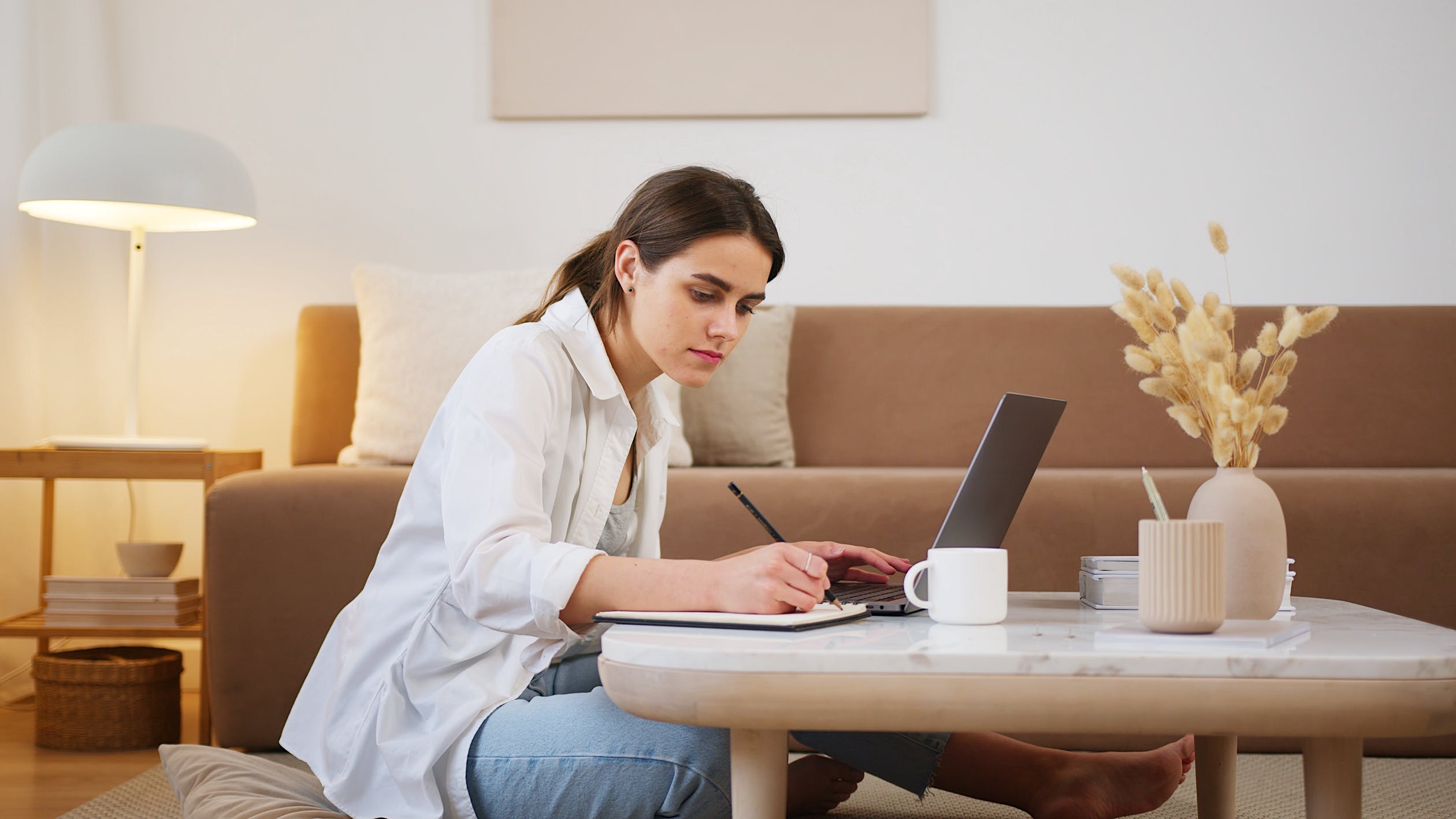 Woman in living room studying from a laptop, taking notes in an A4 notepad