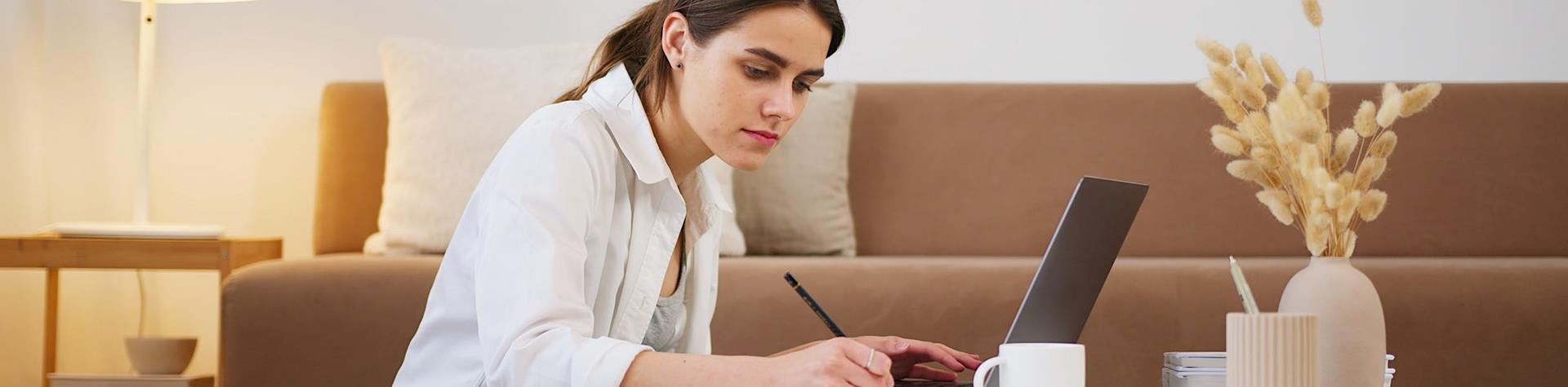 Woman in living room studying from a laptop, taking notes in an A4 notepad